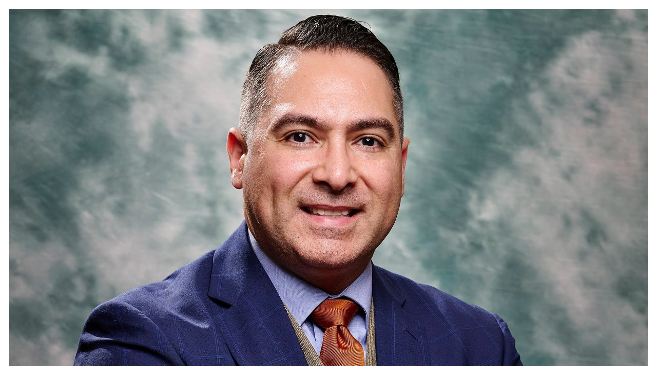 Nestor Torres, a Latino man with short black hair, shown from the shoulders up. H is wearing a dark blue suit with a red tie, and is set behind a blue-white background. He is facing the viewer and is smiling.