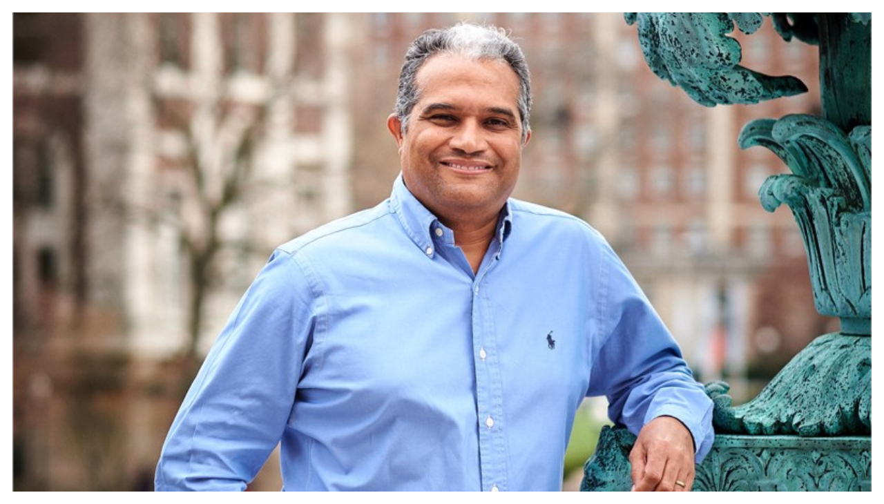 Feniosky Pena-Mora, a Dominican man with short gray hair, shown from the waist up. He is wearing a blue button up shirt, while leaning his arm against a bronze statue. Behind him buildings are visible in the background.