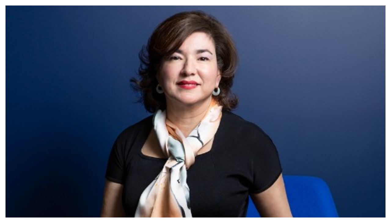 Rebecca Martinez O'Mara, a woman wearing a black shirt with a beige scarf. She is backlit with a dark blue screen behind her.