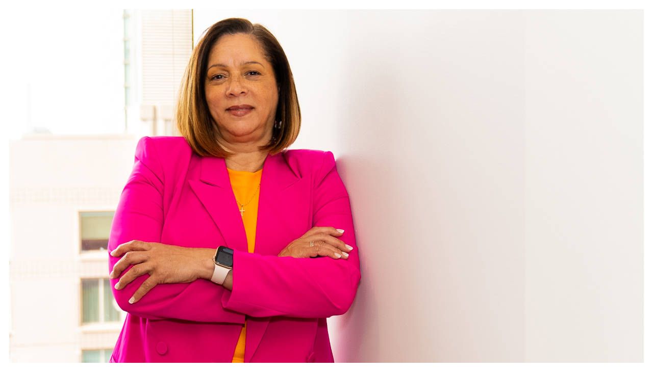 Sandra Carter, a woman wearing a pink business suit leans against a white wall with her hands crossed over her chest.