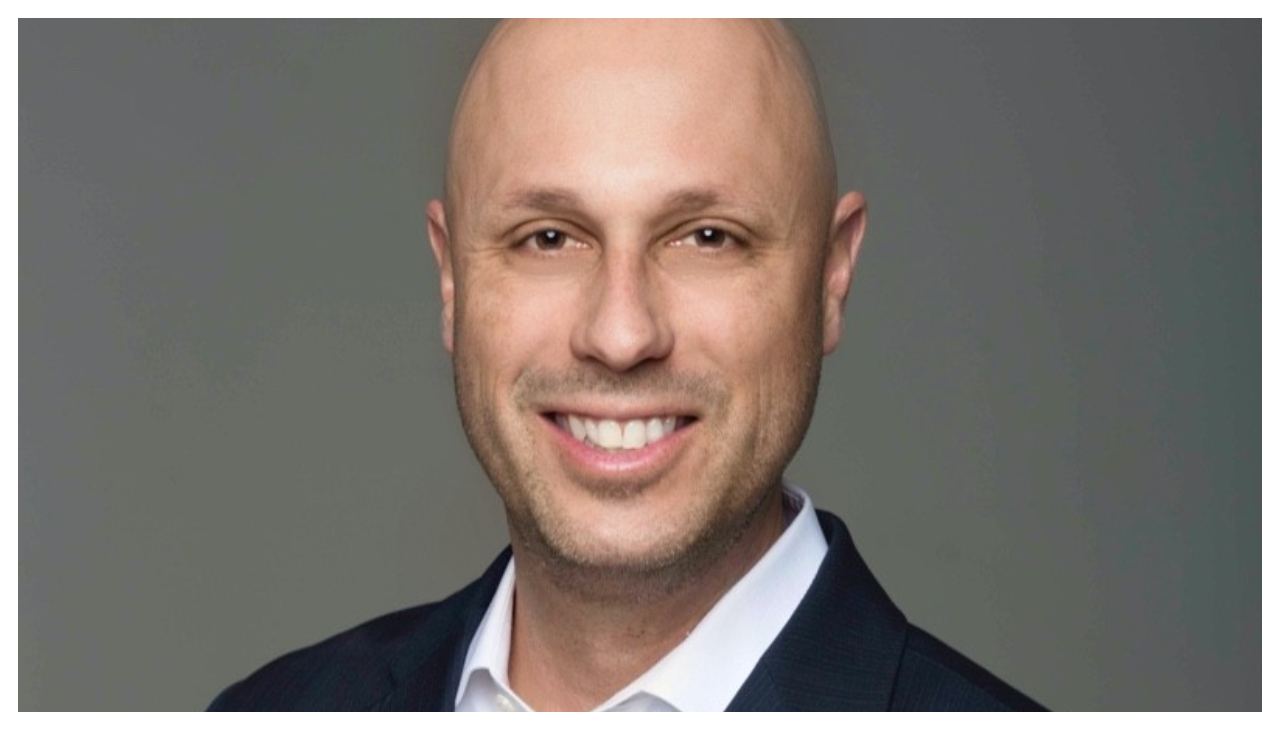 Peter Gonzalez, a Latino man shown from the shoulders up, against a gray background. He is bald, wearing a black suit, and is facing the viewer while smiling.