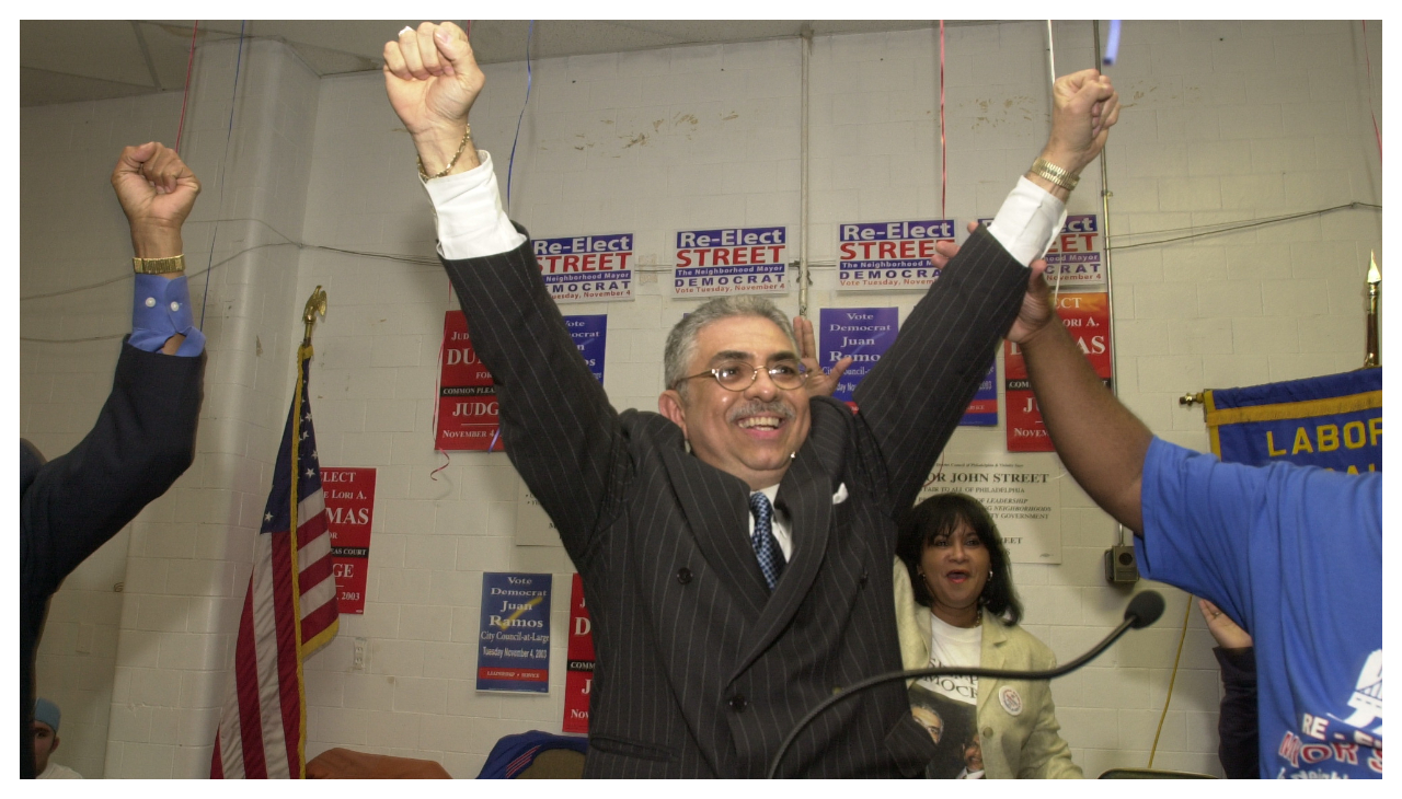 Juan Ramos, a Puerto Rican man, stands before a podium with his arms raised. One is held up by another person, who is also cheering beside several other people out of frame with their arms raised as well.