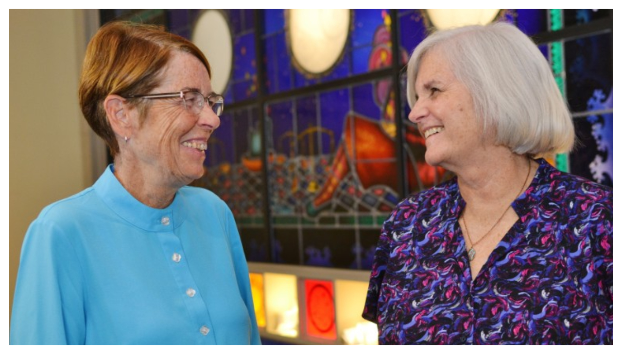 Sister Mary Scullion and Joan Dawson McConnon, speaking to one another. Scullion is an elderly white woman with red hair and a blue button shirt, while McConnon has white hair and a purple button up shirt.