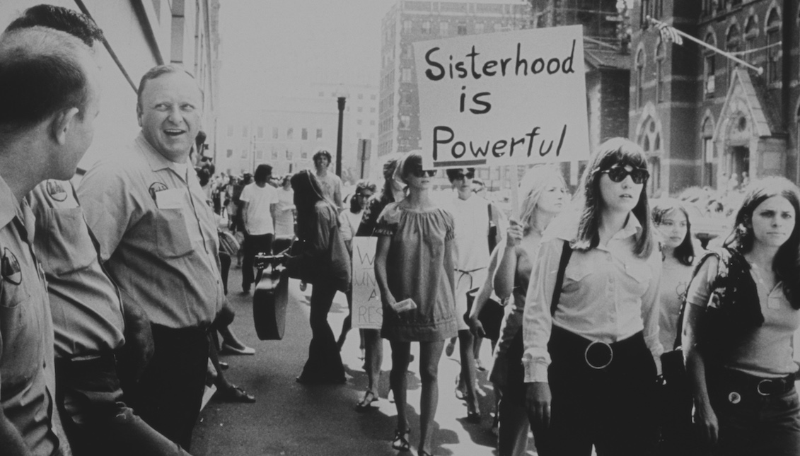 Women marching under a 'Sisterhood is Powerful' placard in the United States, while a group of male workers watch from the left, circa 1975. (Photo by Michael Ochs Archives/Getty Images)