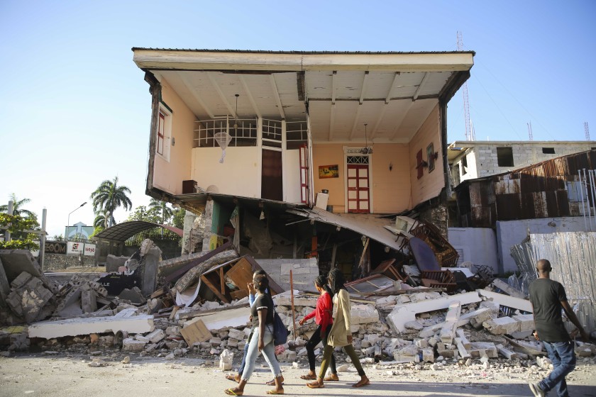 People walk past a home destroyed by the earthquake in Les Cayes, Haiti, Saturday, Aug. 14, 2021. Photo Credit: Joseph Odelyn/AP Photo