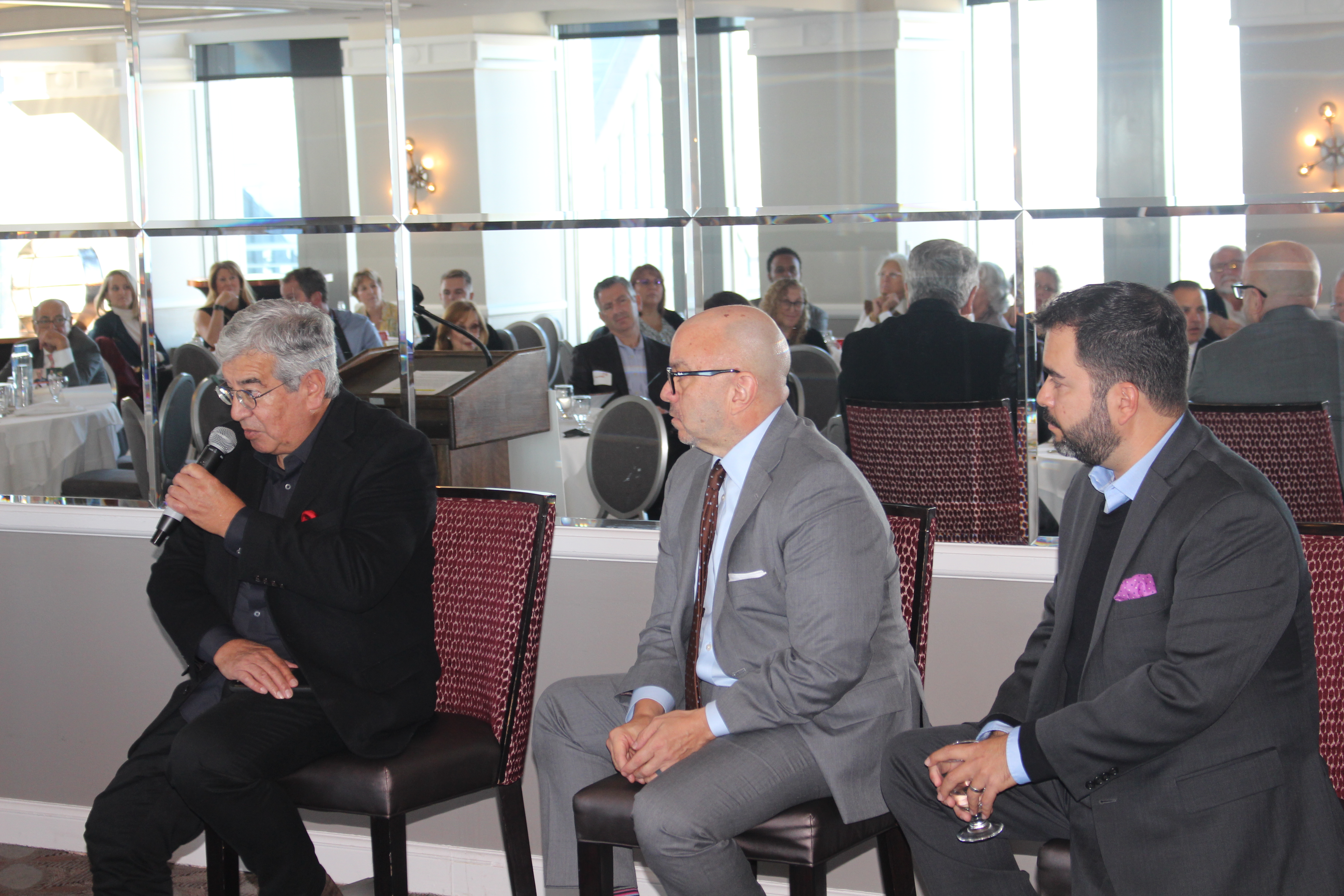 Panelists Fernando Treviño, Larry Gonzalez, and Fernando Mendez sitting on chairs. Mendez is speaking into the microphone.