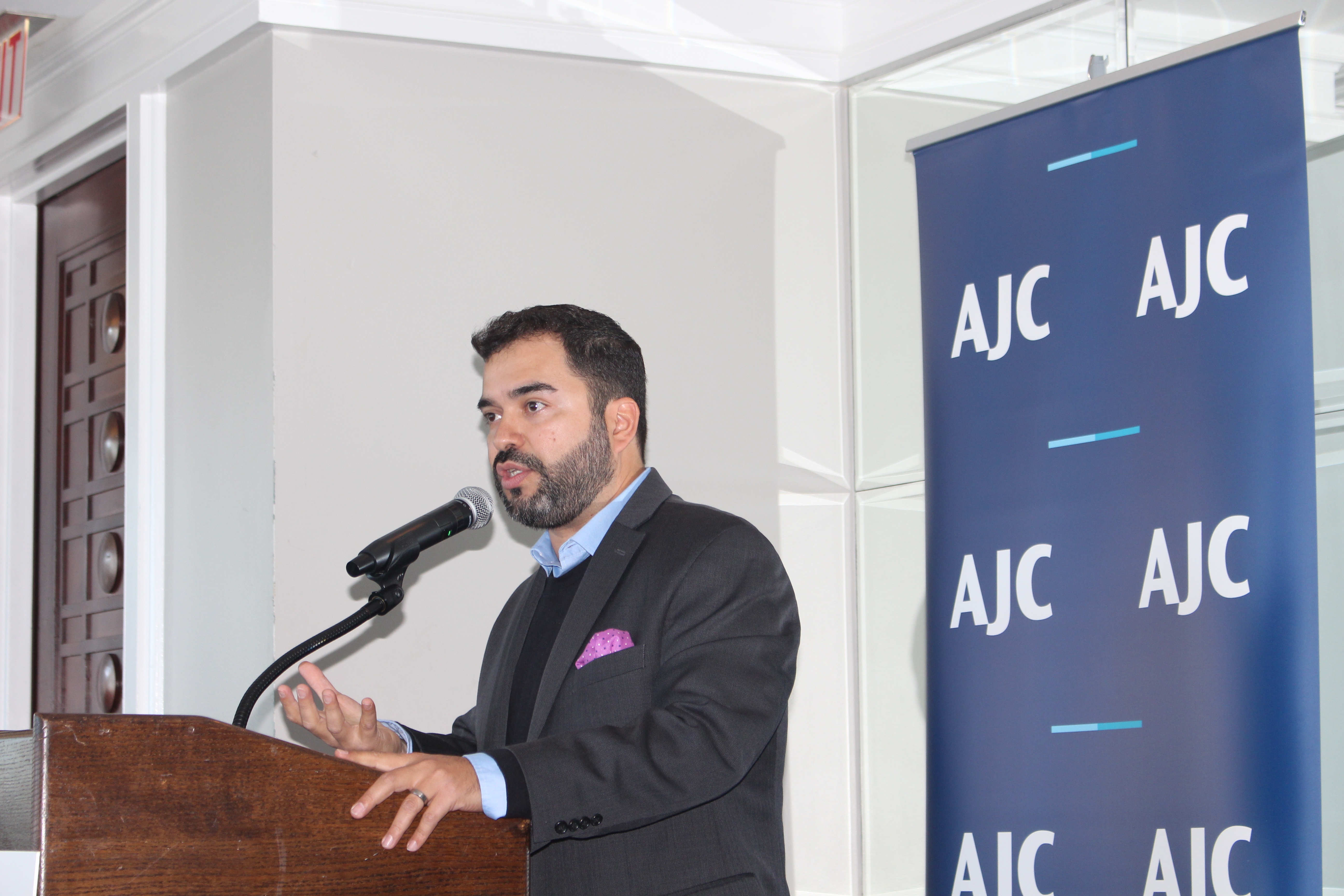 Fernando Treviño, a Latino man in a black suit standing at a podium, speaking into the microphone. Behind him is a photo background with the acronym AJC on it.